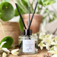 College Green Cotswold Bluebell reed diffuser in a circular heavy glass jar with three thick black reeds. On a wooden counter with flower petals and plants.