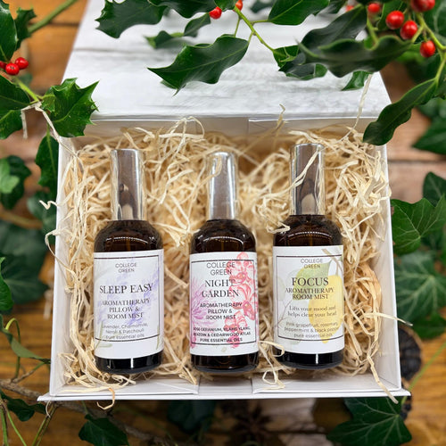 Three room mist bottles from Colleen Green's 'The Holistic Garden' set, displayed in a white gift box filled with shredded paper. The bottles are nestled in natural fiber packing material, framed by green holly leaves and red berries on a rustic wooden surface.