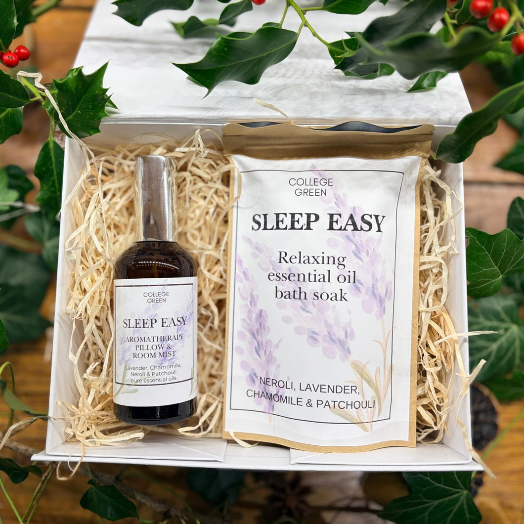 A gift set from College Green featuring a small amber bottle of aromatherapy essential oil and a packet of 'Sleep Easy' relaxing bath soak, nestled in shredded paper inside an open white box with green leaves and berries framing the top.