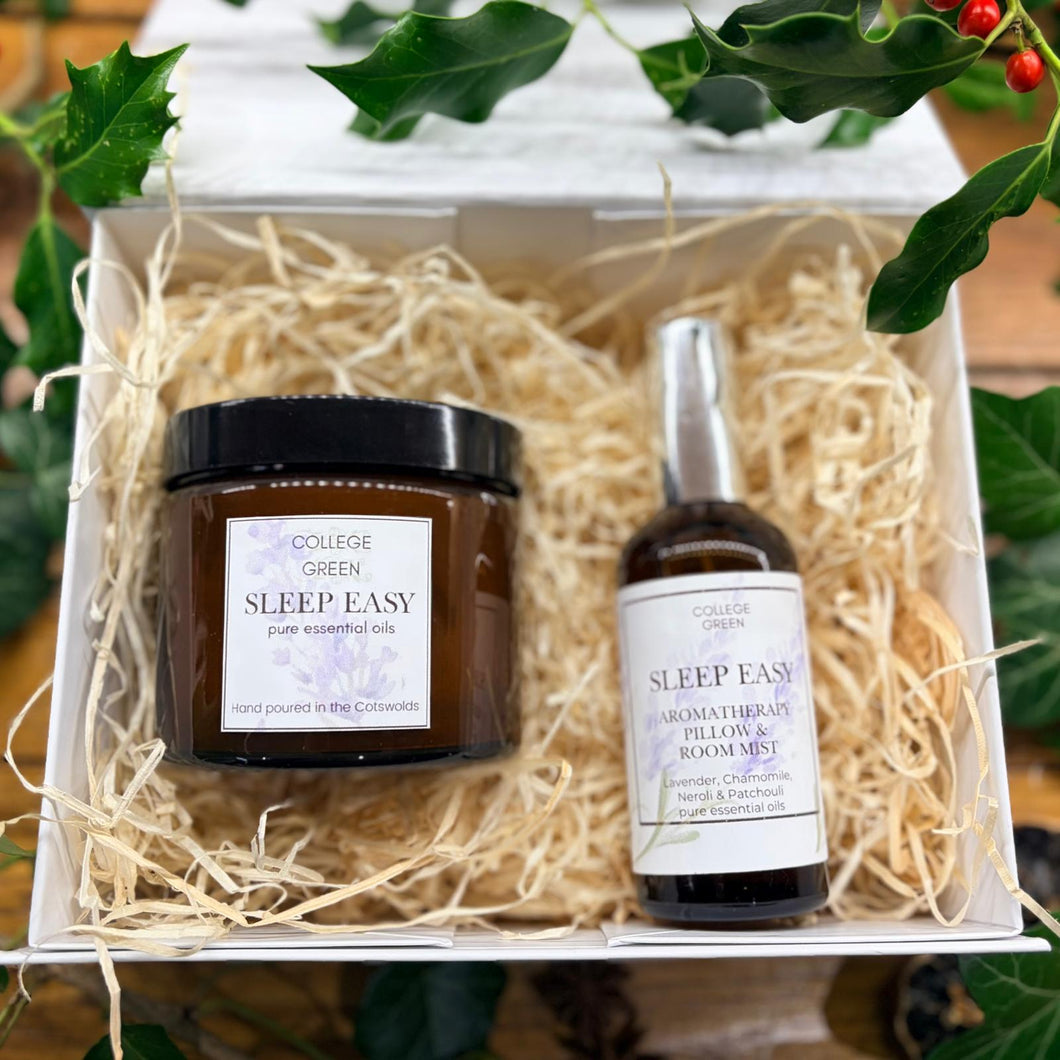 A gift set of two College Green 'Sleep Easy' products in a white box filled with shredded paper, surrounded by holly leaves. The set includes a 'Pure Essential Oils' jar and an 'Aromatherapy Pillow & Room Mist' bottle.