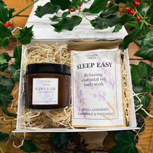 A Sleep Easy essential oil candle in an amber glass jar and a packet of Sleep Easy relaxing essential oil bath soak nestled in a gift box surrounded by greenery