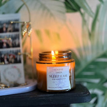 A lit amber glass jar candle labeled 'College Green Sleep Easy' sits on a dark tabletop next to a white jewelry stand with gold-tone rings and earrings, with a blurred background of green leaves and warm ambient light.