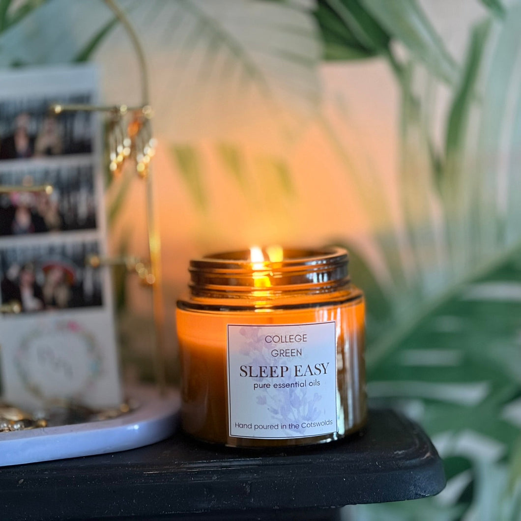 A lit amber glass jar candle labeled 'College Green Sleep Easy' sits on a dark tabletop next to a white jewelry stand with gold-tone rings and earrings, with a blurred background of green leaves and warm ambient light.
