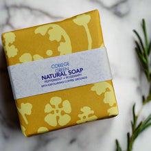 Wrapped bar of Peppermint & Rosemary Coffee Scrub soap on a wooden soap dish