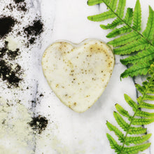 An unwrapped heart shaped bar of Peppermint and Rosemary soap on a white marble surface with coffee grounds and green fern leaves